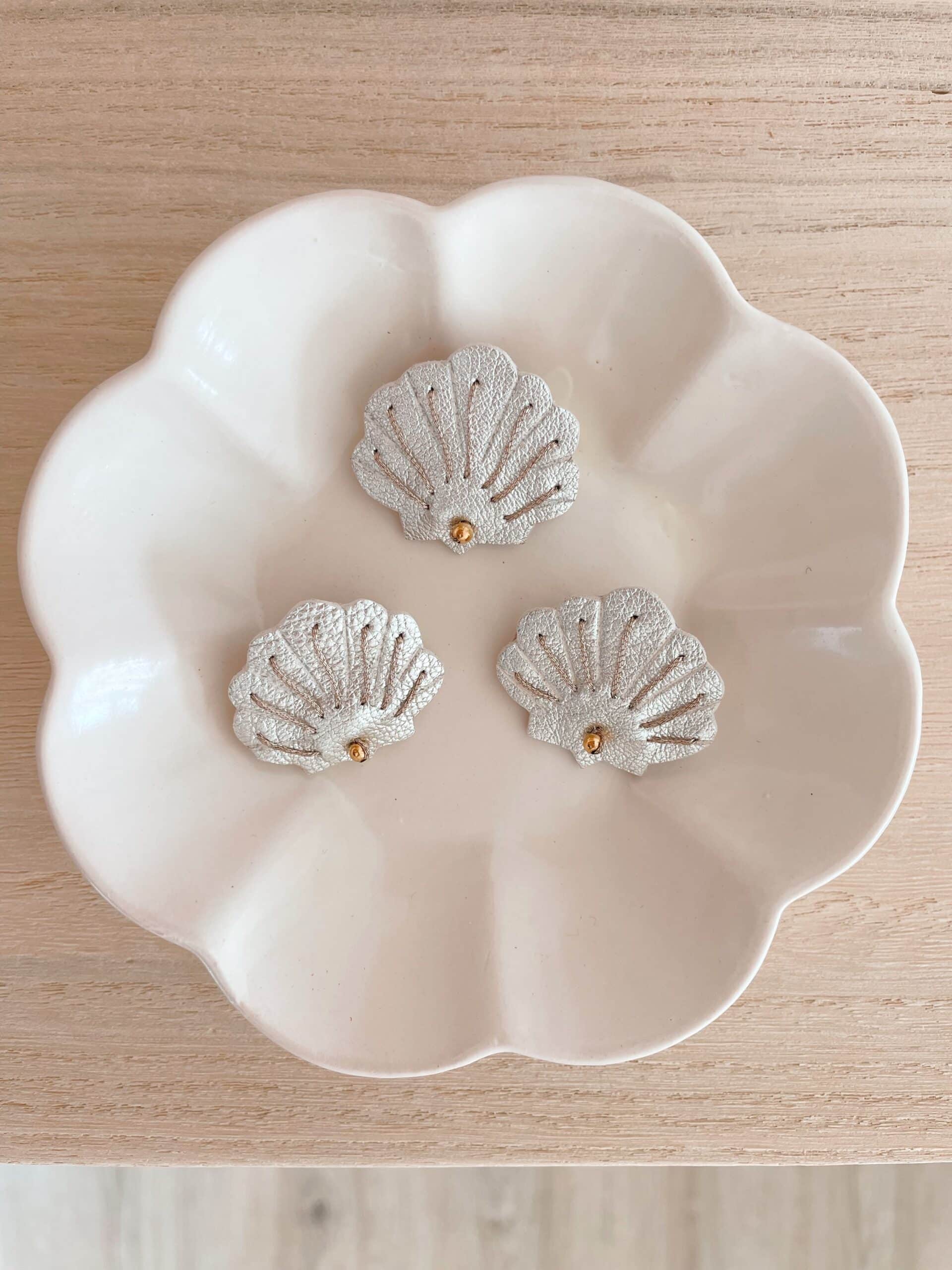 Trois broches coquille Saint-Jacques en cuir surcyclé argenté posées dans une coupelle blanche