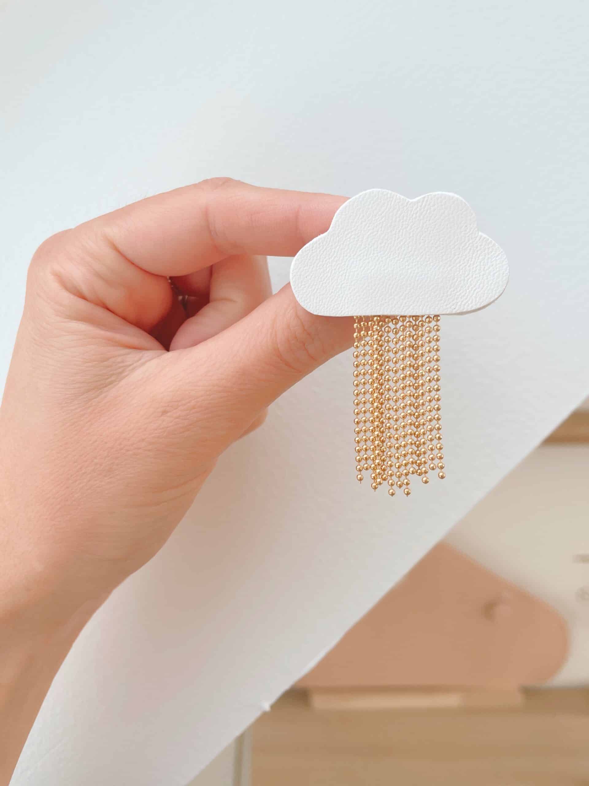 Broche nuage L'Ondée cuir tenue en main — détail pluie dorée © Du vent dans mes valises Broche nuage L'Ondée en cuir surcyclé blanc tenue en main, pluie de chaînes dorées déployée, fond blanc