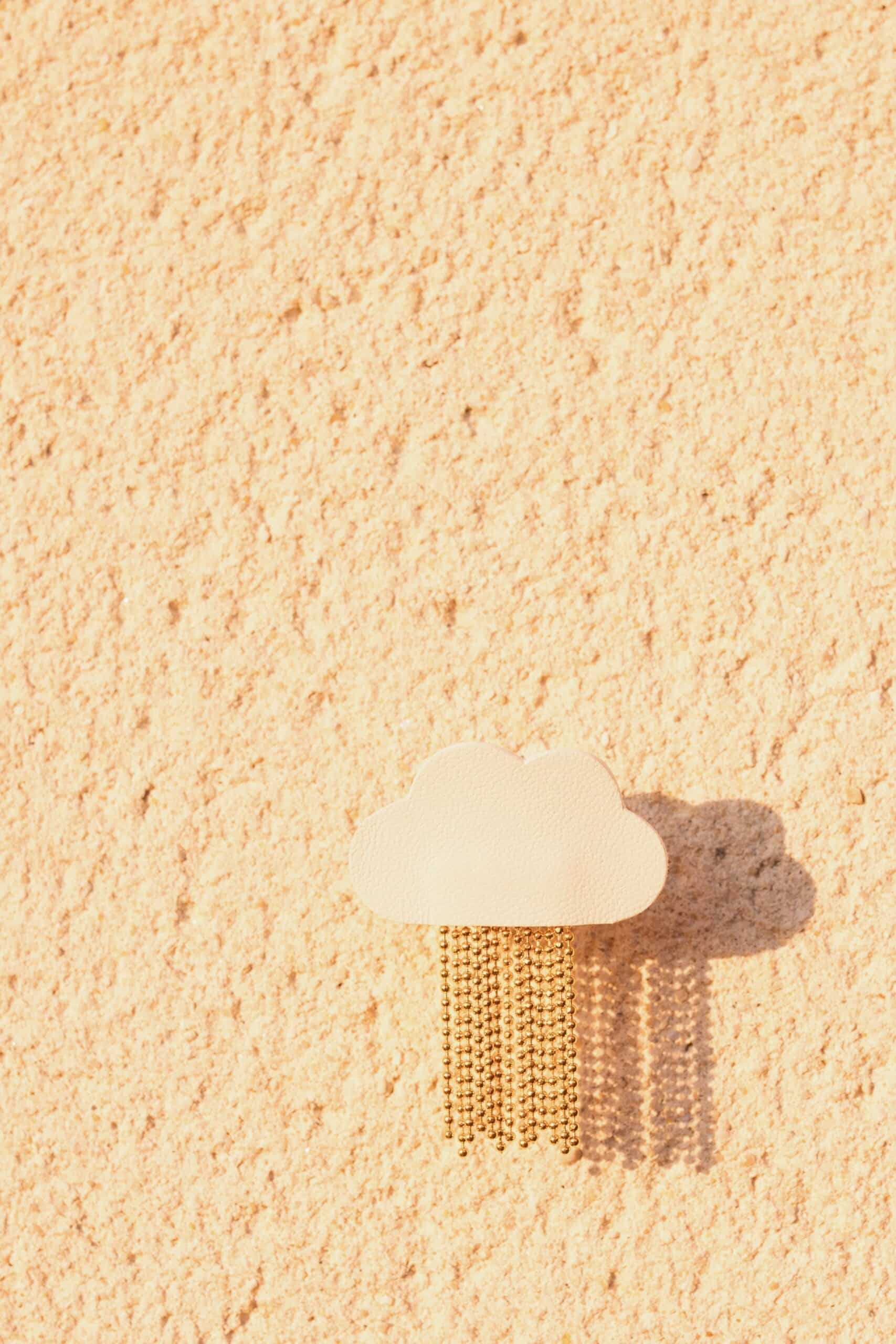 Broche nuage L'Ondée cuir — sur le sable © Du vent dans mes valises Broche nuage L'Ondée en cuir surcyclé blanc posée sur le sable, ombre portée, pluie dorée