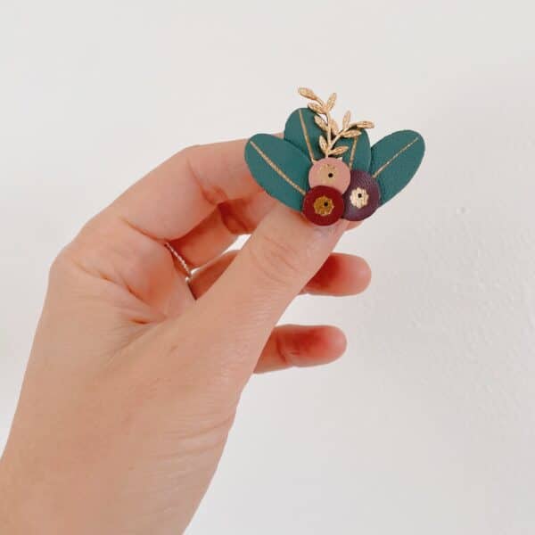 Broche baies en cuir surcyclé tenue en main, baies rose, bordeaux et prune sur feuilles vert sapin dorées à l'or fin