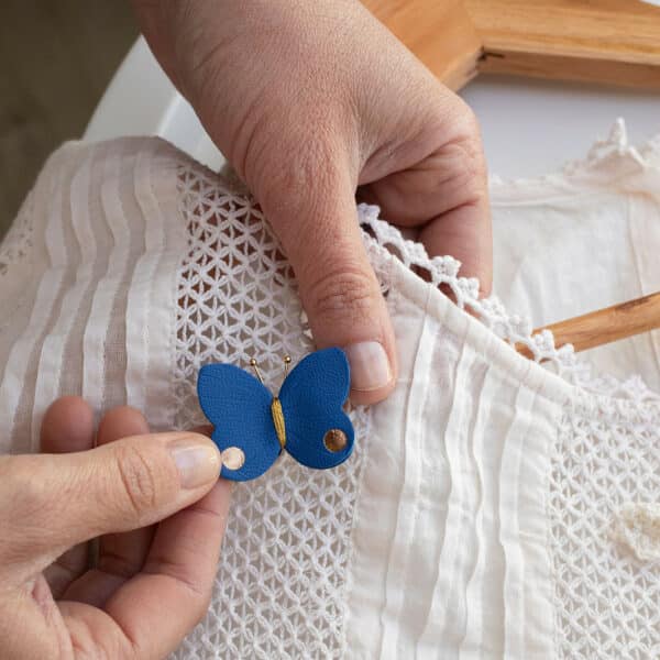 papillon-cuir-bleu-cobalt-dore-tenue-main-du-vent-dans-mes-valises.jpg Alt : Broche papillon en cuir surcyclé bleu cobalt et détails dorés tenue en main sur tissu blanc brodé