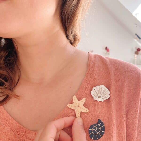 Main épinglant une broche étoile de mer dorée sur t-shirt rose, avec coquille et ammonite