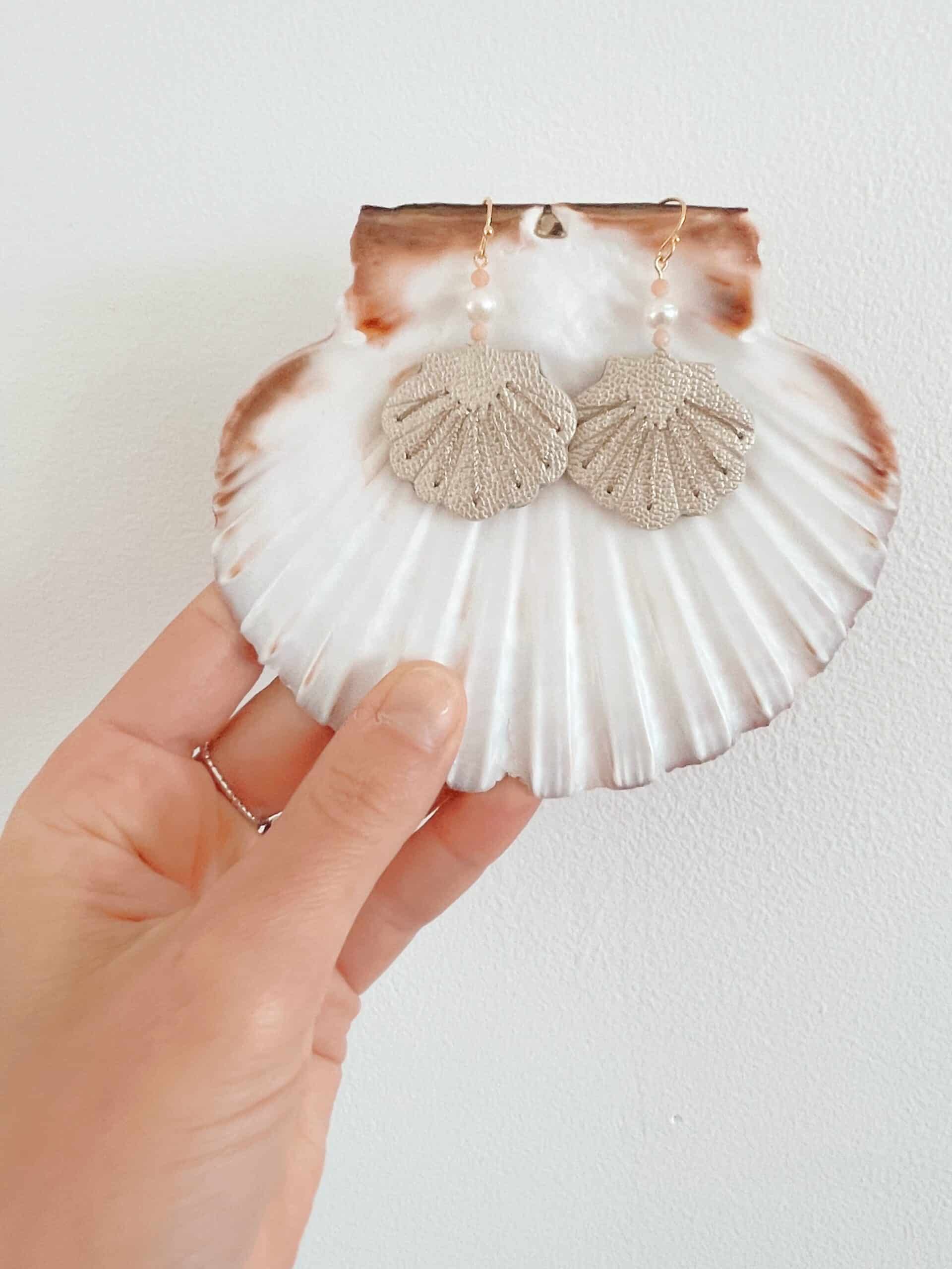 Boucles d'oreilles coquille cuir — sur coquille tenue en main © Du vent dans mes valises Boucles d'oreilles coquille en cuir nacré posées sur une grande coquille Saint-Jacques naturelle tenue en main, fond blanc