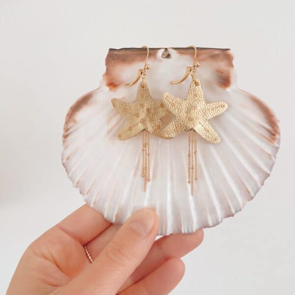 Boucles d'oreilles étoile de mer en cuir doré posées sur une grande coquille naturelle, boîte Du vent dans mes valises en arrière-plan