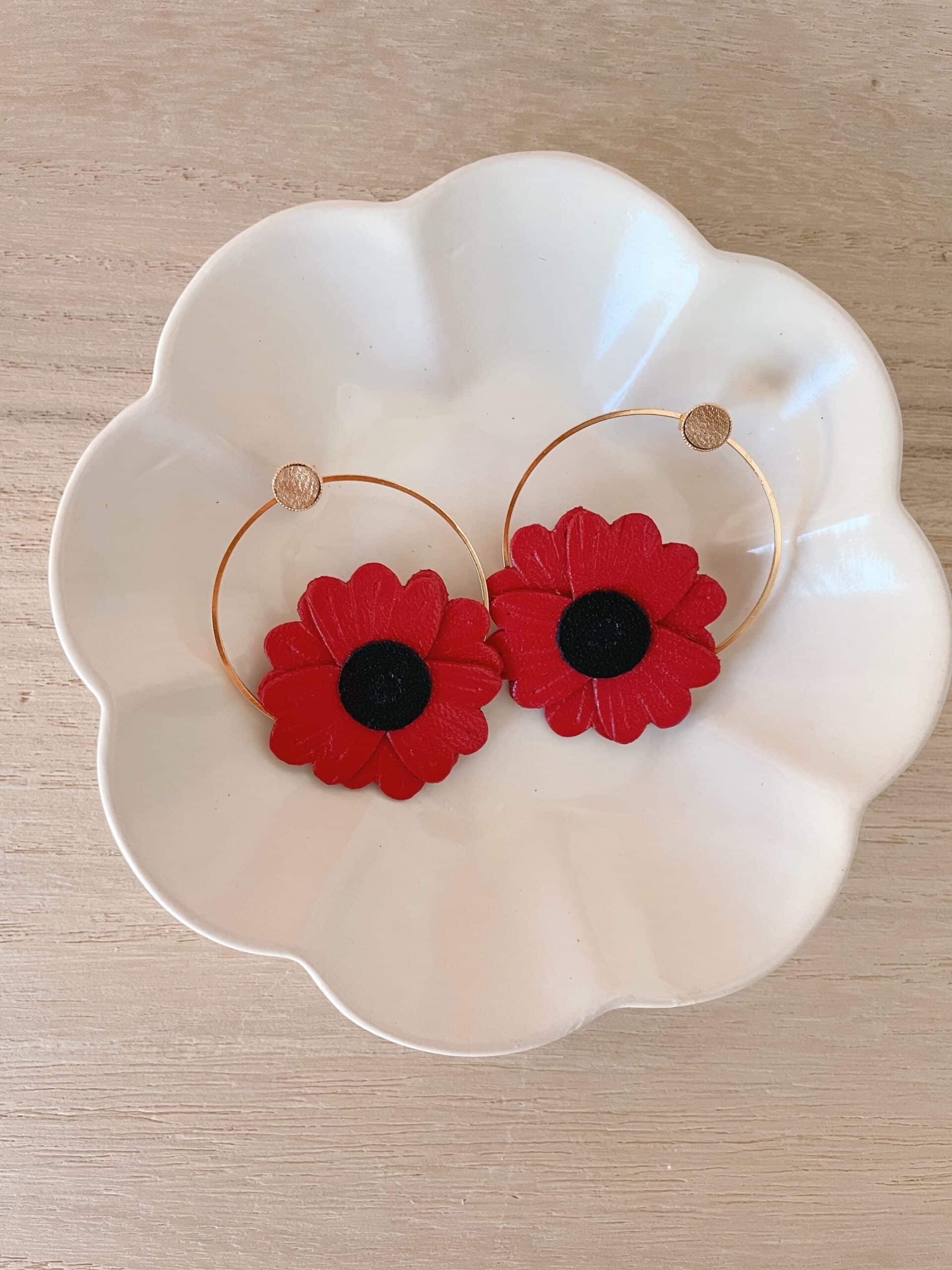 Boucles d'oreilles coquelicot cuir rouge — image à la une © Du vent dans mes valises Paire de boucles d'oreilles coquelicot en cuir surcyclé rouge, cœur noir, sur créoles dorées, posées dans une coupelle blanche