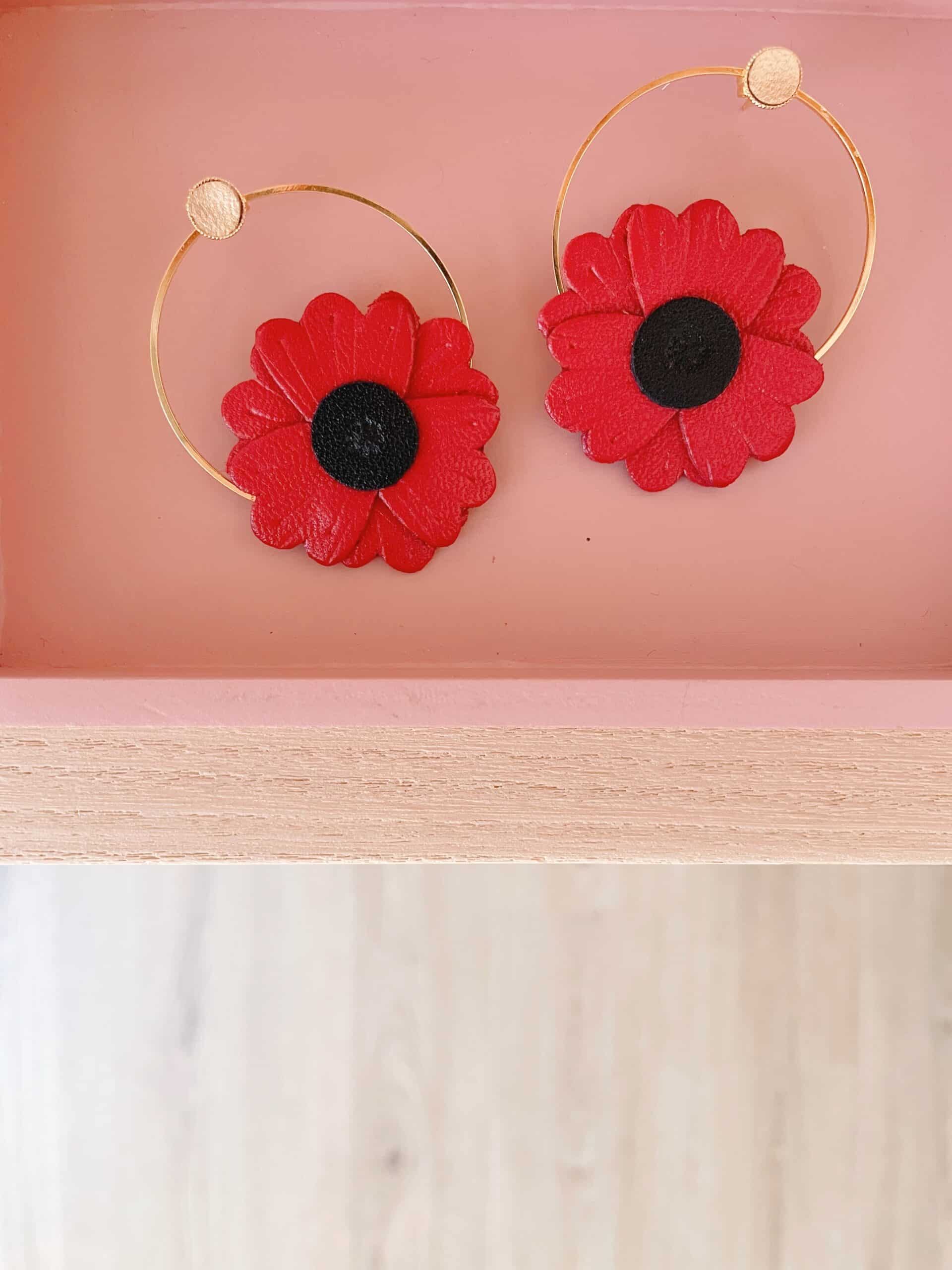 Boucles d'oreilles coquelicot cuir rouge — cadre rose © Du vent dans mes valises Paire de boucles d'oreilles coquelicot en cuir rouge et créoles dorées posées dans un cadre rose