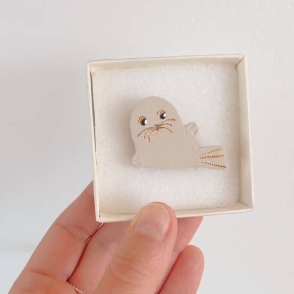 Broche phoque Selkie en cuir surcyclé blanc cassé tenue en main dans son écrin