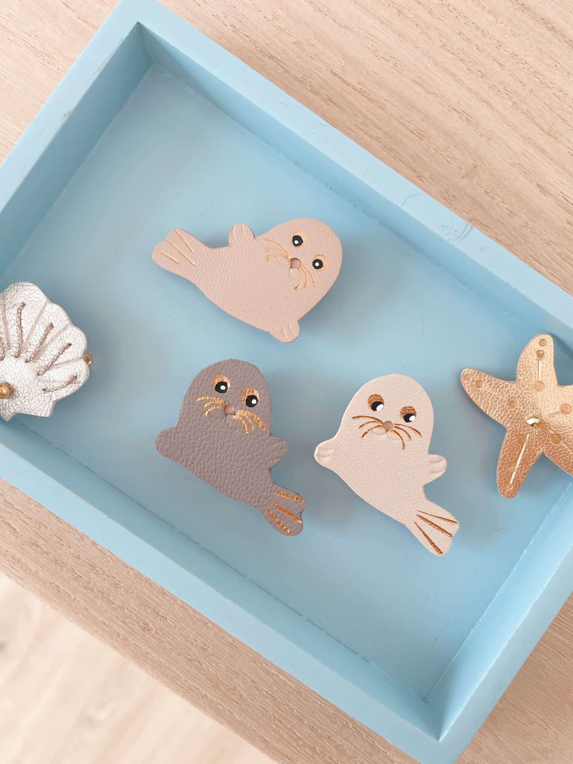 Broche phoque Selkie cuir — image à la une © Du vent dans mes valises Broches phoque Selkie en cuir surcyclé — gris bleuté, beige rosé et blanc ivoire — sur plateau bleu