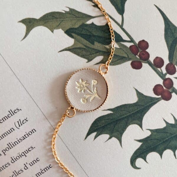 Bracelet médaillon en cuir beige doré à l’or fin d’un edelweiss, posé sur une planche botanique de houx